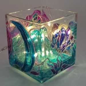 Può includere: Un portacandele quadrato in vetro con disegni astratti dipinti a mano nei toni del verde acqua, viola e rosa. L'interno è illuminato da piccole luci a LED bianche calde, che creano una luce soffusa. Un elemento decorativo per la casa.