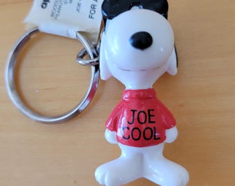 May include: A Joe Cool keychain featuring a white Snoopy figure wearing a red shirt with "JOE COOL" printed on it. The keychain includes a metal ring and a tag with the "applause" logo and text that reads "FOR AGES OVER 3".