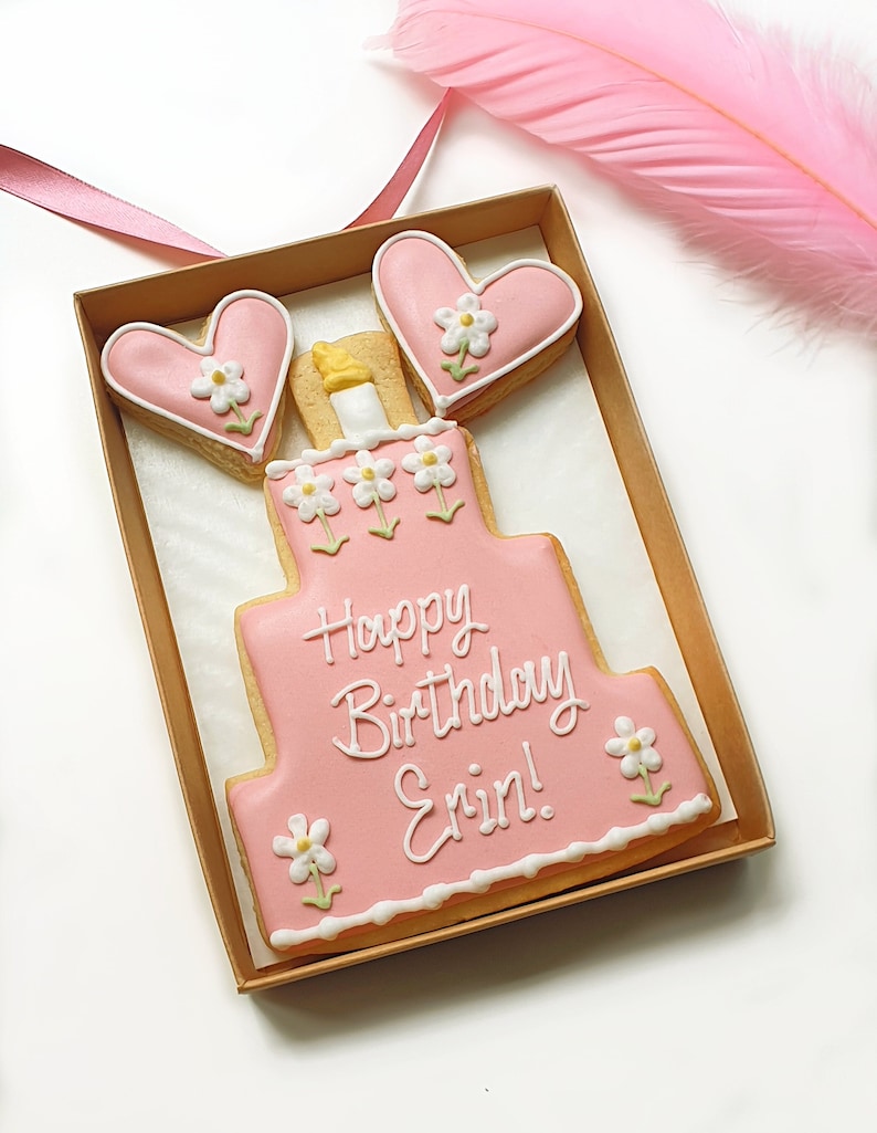 May include: A pink and white decorated sugar cookie in the shape of a birthday cake with the words "Happy Birthday Erin!"  The cookie is in a brown box with two pink heart-shaped cookies on top.