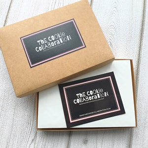 May include: A brown cardboard box with a white label that says "The Cookie Collaboration" in black text. The box is open and there is a white card inside with a black label that says "The Cookie Collaboration" in white text. The card also has the website address "www.thecookiecollaboration.co.uk" printed on it.