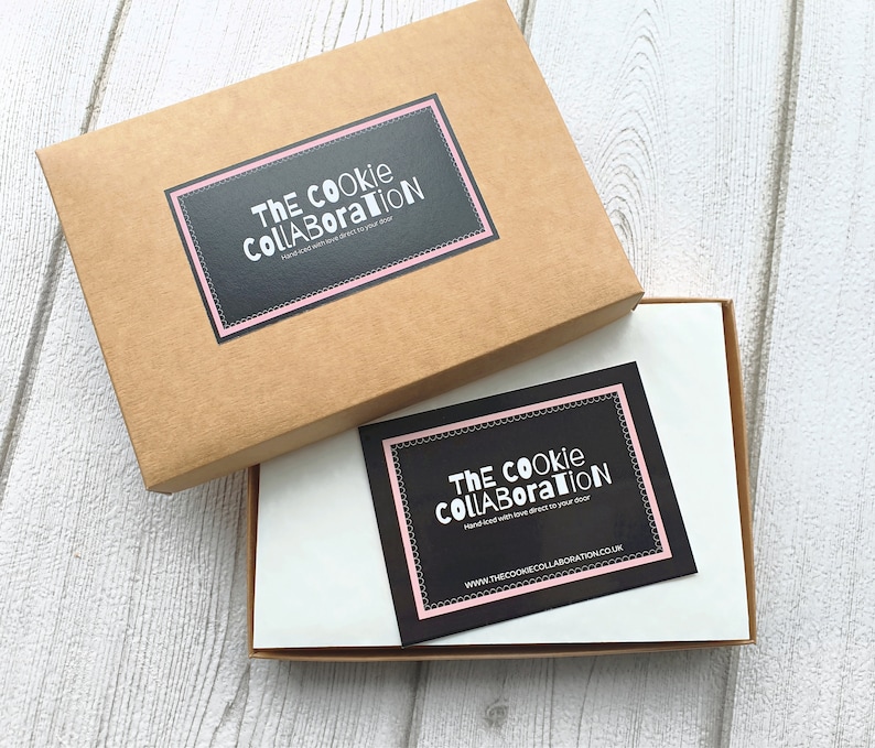 May include: A brown cardboard box with a white label that says "The Cookie Collaboration" in black text. The box is open and there is a white card inside with a black label that says "The Cookie Collaboration" in white text. The card also has the website address "www.thecookiecollaboration.co.uk" printed on it.