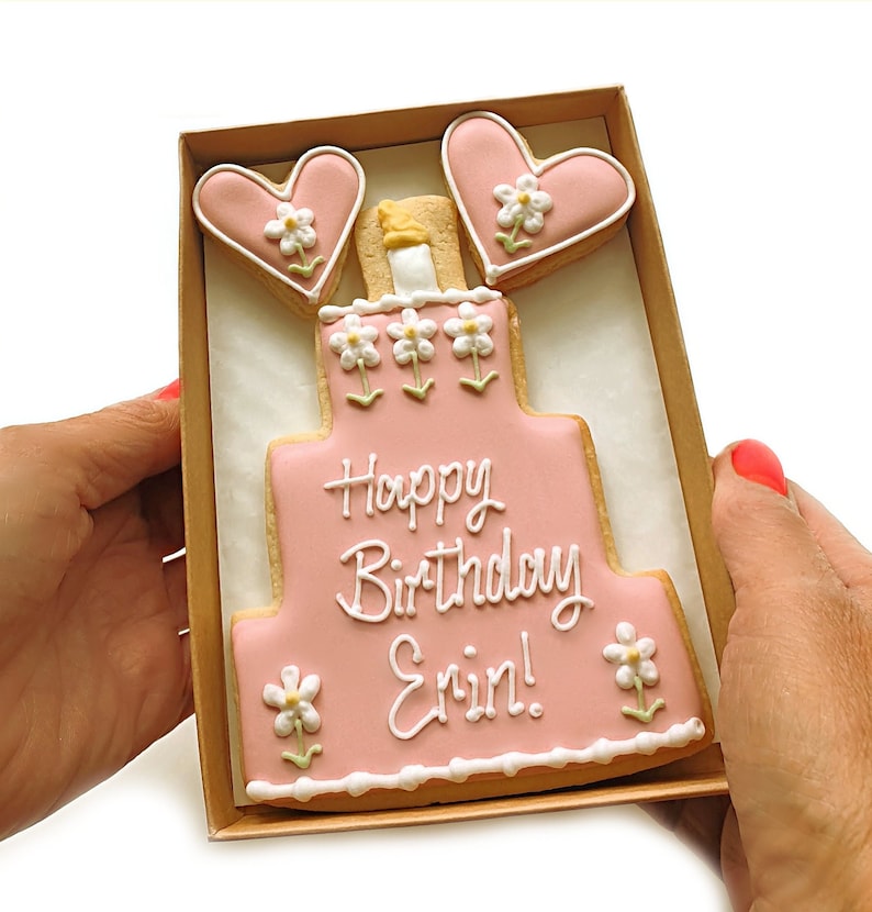 May include: A pink and white decorated sugar cookie in the shape of a cake with the words "Happy Birthday Erin!"  The cookie is in a brown cardboard box with two pink heart-shaped cookies with white flowers on top.