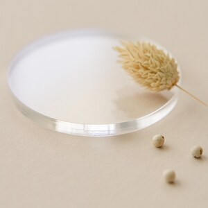 May include: A clear glass round dish with a dried flower on top. There are three white peppercorns on the beige surface.