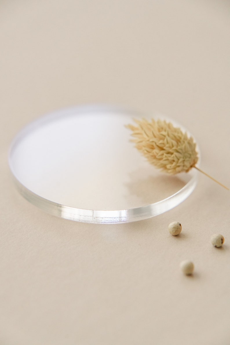 May include: A clear glass round dish with a dried flower on top. There are three white peppercorns on the beige surface.