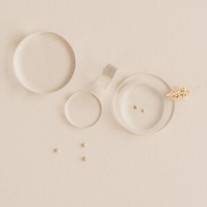 May include: Three clear glass dishes of varying sizes, two with small gold beads inside, and one with a dried wheat stalk. Three small white beads are scattered on the beige surface.