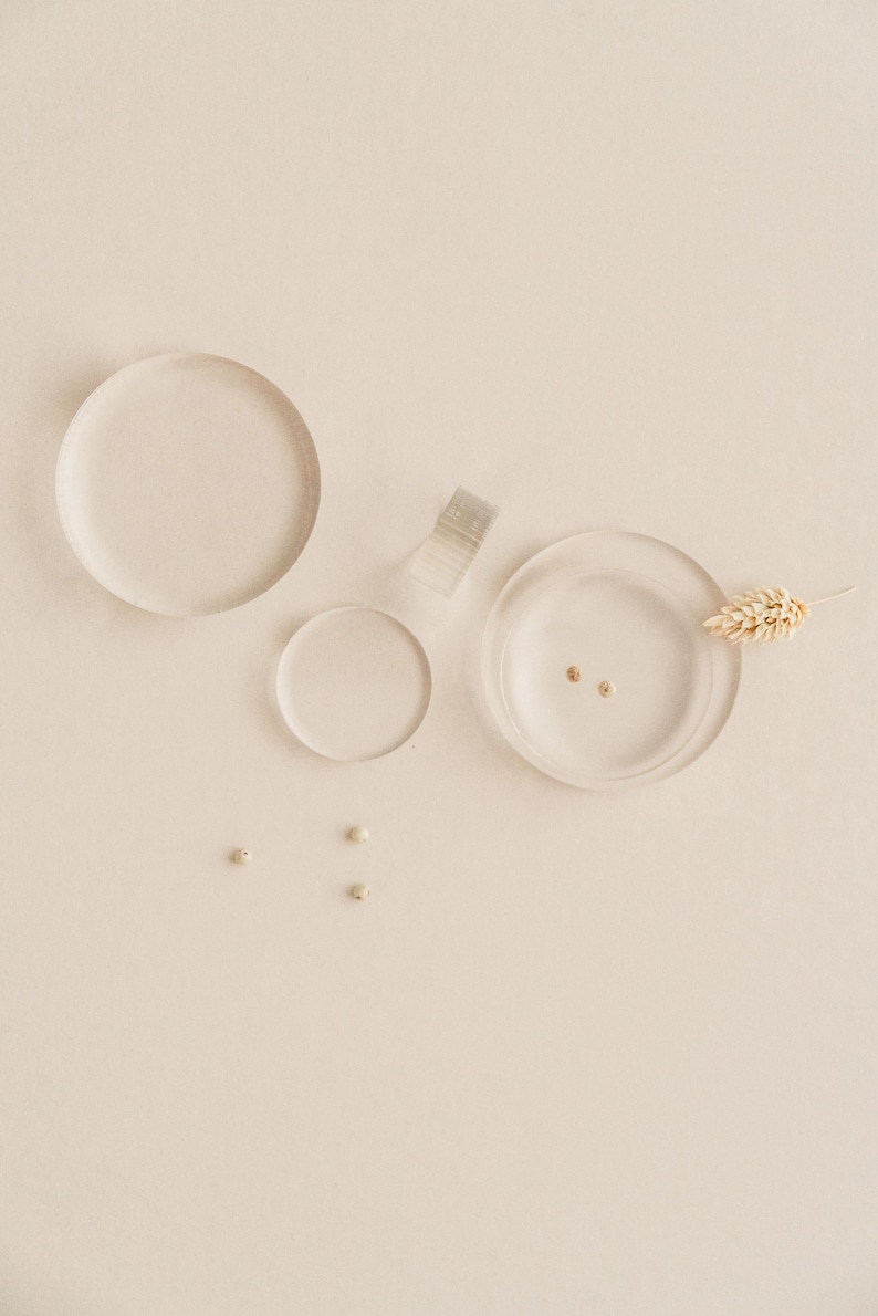 May include: Three clear glass dishes of varying sizes, two with small gold beads inside, and one with a dried wheat stalk. Three small white beads are scattered on the beige surface.