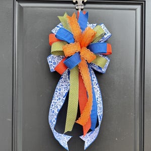 May include: A decorative door bow featuring a mix of blue, orange, green, and white ribbons. The bow has a floral pattern on the blue ribbons and is accented with orange lace trim. The bow is attached to a black door.