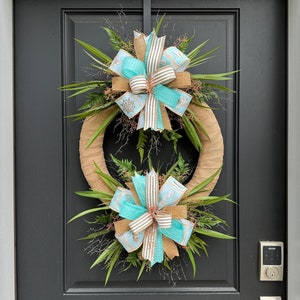 May include: A double-layered burlap wreath with two turquoise and brown bows. The wreath is decorated with green foliage and is hanging on a black door.