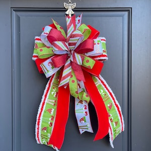 May include: A red, green, and white Christmas bow with a festive elf design. The bow is tied with a red velvet ribbon and is ready to be hung on a door.