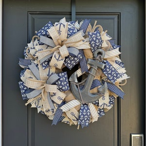 May include: A navy blue and tan wreath with a large wooden anchor in the center. The wreath is decorated with white anchors and nautical rope.
