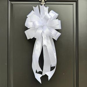 May include: A large, white decorative bow made of satin ribbon, attached to a black door. The bow has multiple loops and long, flowing streamers. The bow is centered on the door, creating a classic and elegant look.