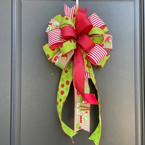 May include: A Christmas door bow with red, white, and green ribbon. The bow has a red glitter ribbon in the center and is decorated with a green and white polka dot ribbon, a red and white striped ribbon, and a burlap ribbon with a gnome pattern.