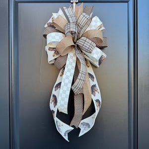 May include: A brown, white, and black horse-themed bow with a checkered pattern and brown polka dots. The bow is tied with a burlap ribbon and is hanging on a black door.
