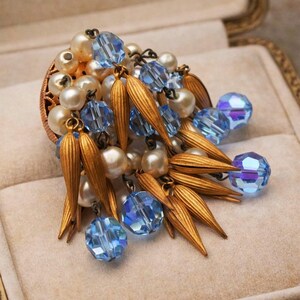 Puede incluir: Broche vintage con perlas de imitación, cristales azules y detalles metálicos dorados. El broche se exhibe en una caja decorativa con forro de terciopelo beige, resaltando su intrincado diseño y paleta de colores.