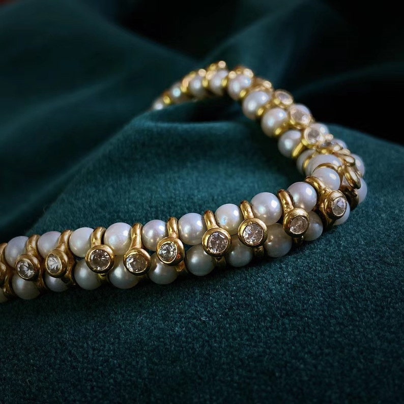 Op de afbeelding: Een dubbele rij parel armband met gouden accenten en heldere edelstenen. De armband ligt op een donkergroene stoffen achtergrond.