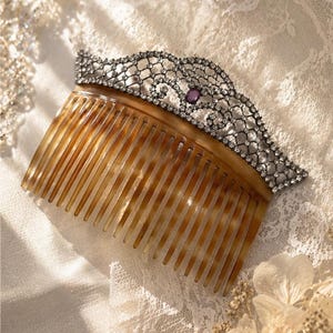 May include: A vintage hair comb with a decorative silver filigree top adorned with clear crystals and a central purple gemstone. The comb's base is a warm, tortoiseshell color, set against a backdrop of delicate lace.