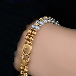 Op de afbeelding: Een gouden armband met een dubbele rij kralen. De ene rij is gemaakt van gouden kralen en de andere rij is gemaakt van witte parels. Een kleine diamant is in het midden van de armband gezet.