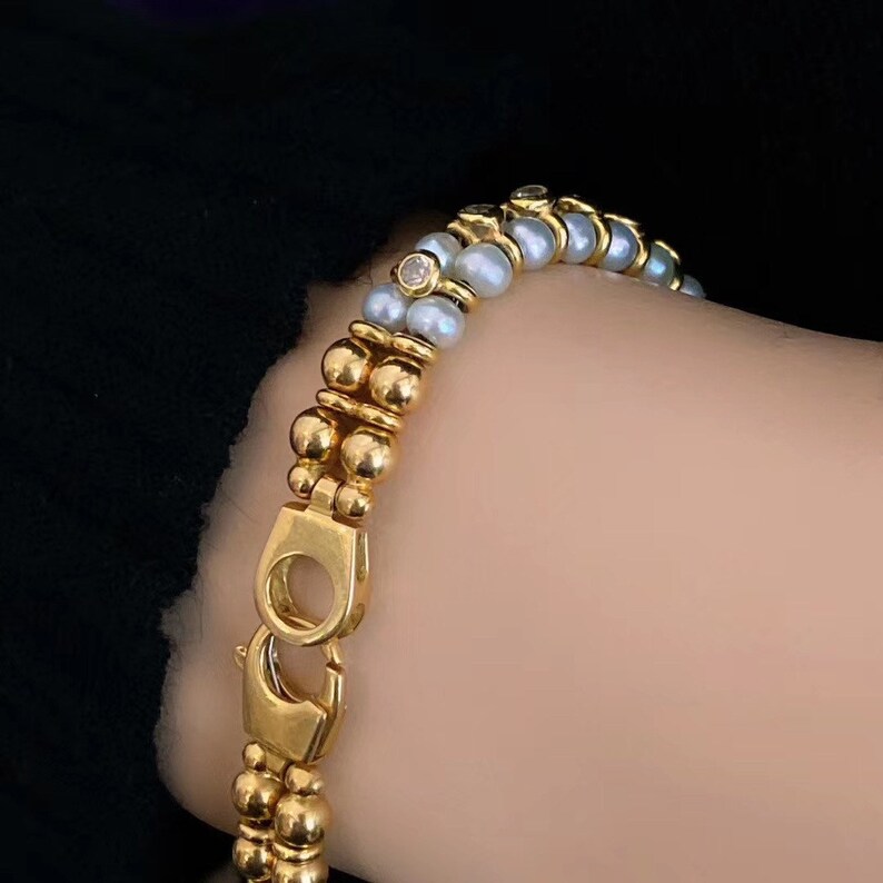 Op de afbeelding: Een gouden armband met een dubbele rij kralen. De ene rij is gemaakt van gouden kralen en de andere rij is gemaakt van witte parels. Een kleine diamant is in het midden van de armband gezet.