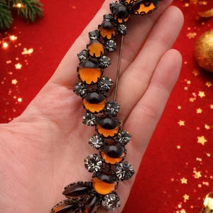 Puede incluir: Broche vintage con diseño de abeja, con detalles en naranja y negro, adornado con brillantes cristales grises. El broche se sostiene en una mano sobre un fondo rojo, con adornos dorados.