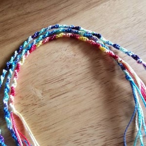 May include: Close-up of three colorful friendship bracelets. Each bracelet is made of twisted threads in various colors, including red, blue, yellow, and white. The bracelets are lying on a wooden surface.