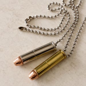 Può includere: Due ciondoli a forma di proiettile attaccati a catenine a palline color argento. Un ciondolo presenta un bossolo color argento lucido con una punta color rame, mentre l'altro ha un bossolo color ottone con una punta color rame abbinata.