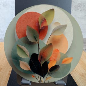May include: A circular decorative plaque featuring a layered botanical design. The artwork displays overlapping circles in shades of sage green, burnt orange, and cream, accented by delicate, translucent leaf silhouettes in dark and light tones.