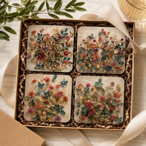 May include: A set of four square coasters arranged in a brown gift box with crinkle paper and a beige ribbon. Each coaster features a textured off-white surface embedded with pressed dried flowers in shades of red, orange, yellow, and teal green.