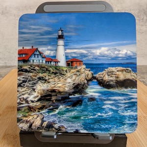 May include: A coaster with a coastal scene featuring a white lighthouse with a black top, red-roofed buildings, and a blue sea. The sky is a bright blue with fluffy white clouds. The image depicts a rocky coastline.