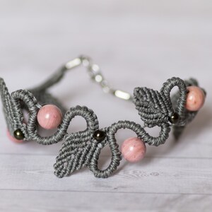 Peut inclure: Un bracelet en macramé gris avec des perles roses et brunes. Le bracelet est réalisé avec une technique de nouage et présente un motif de feuilles.