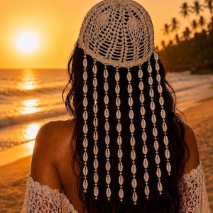 May include: A woman's back is shown, wearing a cream-colored, crocheted head covering with dangling strands of seashells. The setting sun casts a warm glow over the beach and ocean, creating a serene and bohemian aesthetic.
