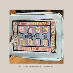May include: A light blue pouch featuring a needlepoint canvas insert with the text BAM! DOT! CRAK! and MAHJONG. The canvas displays colorful mahjong tile illustrations and a floral border. The text STITCHING NOT COMPLETE IN THIS PHOTO is visible.