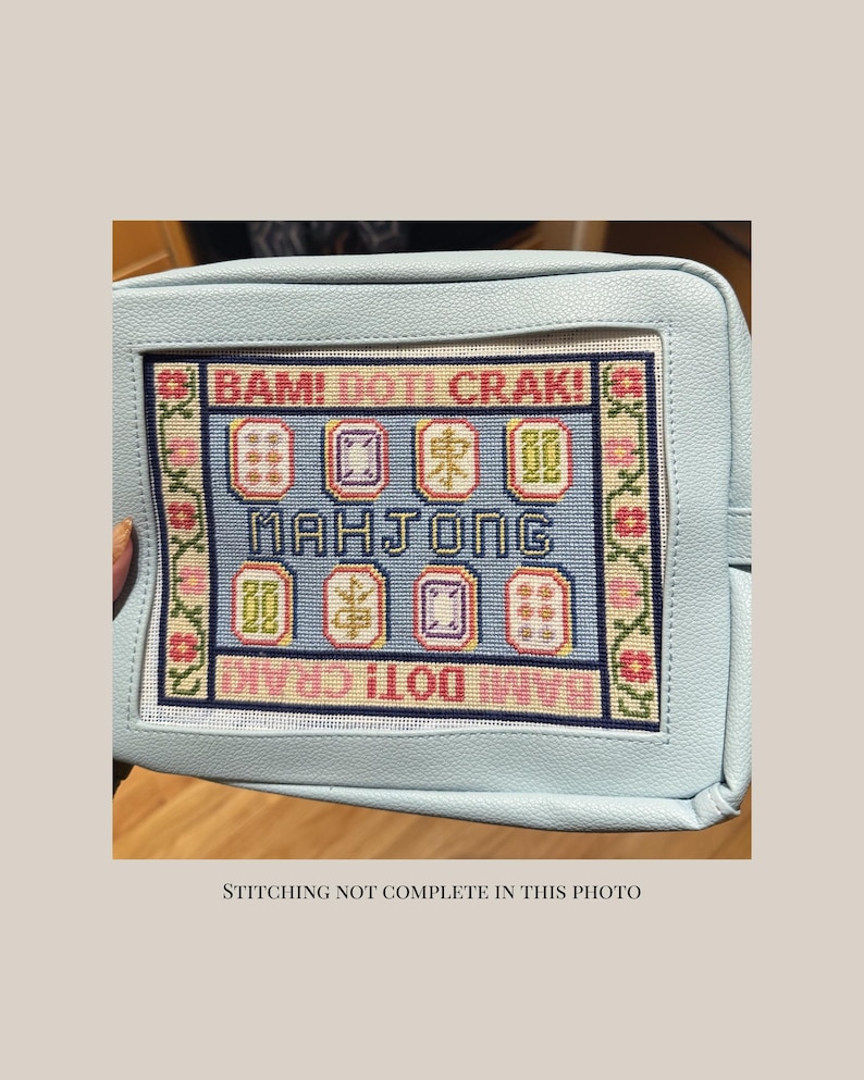 May include: A light blue pouch featuring a needlepoint canvas insert with the text BAM! DOT! CRAK! and MAHJONG. The canvas displays colorful mahjong tile illustrations and a floral border. The text STITCHING NOT COMPLETE IN THIS PHOTO is visible.