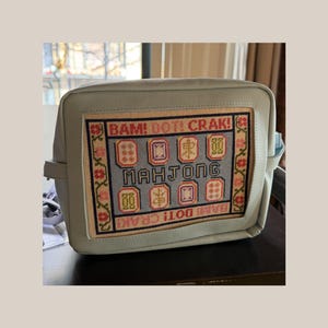 May include: A light blue rectangular bag featuring a central needlepoint panel. The panel displays the text BAM! DOT! CRAK! and MAHJONG, surrounded by colorful embroidered mahjong tiles and a floral border in shades of red, pink, and green.