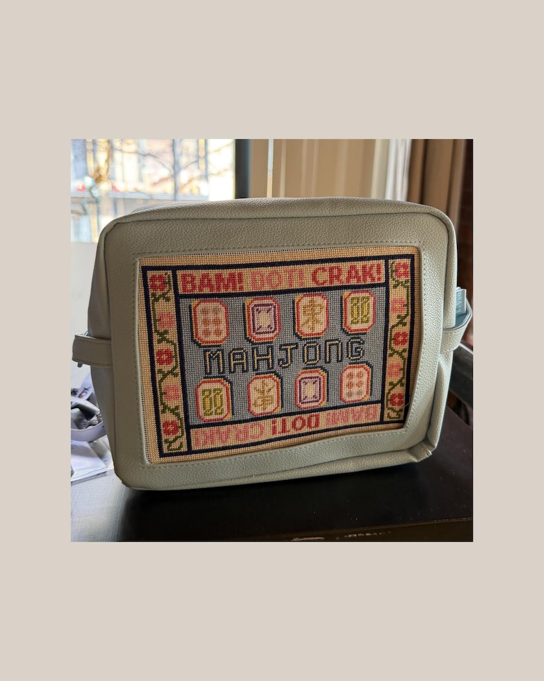 May include: A light blue rectangular bag featuring a central needlepoint panel. The panel displays the text BAM! DOT! CRAK! and MAHJONG, surrounded by colorful embroidered mahjong tiles and a floral border in shades of red, pink, and green.