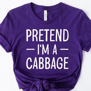 May include: A purple t-shirt with the text "PRETEND - I'M A - CABBAGE" in white. The shirt is knotted at the bottom. The sleeves are cuffed. The shirt is made of a soft material.