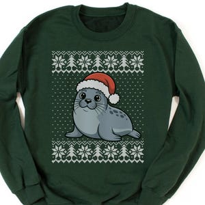 May include: A dark green sweatshirt featuring a cartoon seal wearing a Santa hat. The seal is gray with black spots and has a smiling expression. The sweatshirt has a white snowflake and tree pattern around the seal.