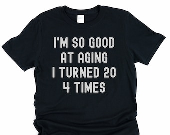 以下が含まれることがあります： 「I'm so good at aging I turned 20 4 times」という白い文字が書かれた黒いTシャツ。