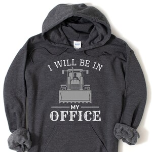 May include: A dark grey hooded sweatshirt with the text "I will be in my office" and a grey silhouette of a tractor with a person in the driver's seat.