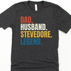 Puede incluir: Camiseta gris oscuro con las palabras "DAD. HUSBAND. STEVEDORE. LEGEND." en letras de bloque rojas, blancas, amarillas y azules. La camiseta es de cuello redondo y está hecha de un material suave.