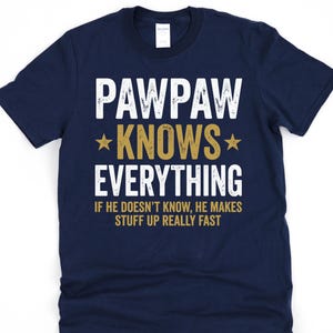 May include: Navy blue t-shirt with the text "PAWPAW KNOWS EVERYTHING" in white and gold lettering. The shirt also includes the phrase "IF HE DOESN'T KNOW, HE MAKES STUFF UP REALLY FAST". A casual tee for a grandparent.