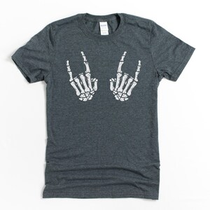 May include: A dark heather gray t-shirt with a white skeleton hand graphic making the rock and roll hand gesture.