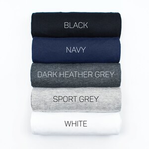 May include: Five rolled up t-shirts in different colors: black, navy, dark heather grey, sport grey, and white. The colors are labeled on each t-shirt.
