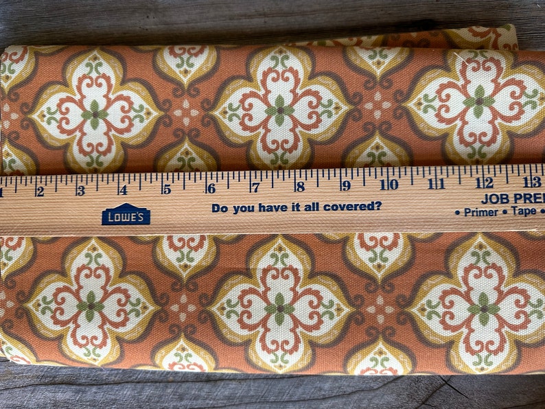 May include: Folded fabric featuring a repeating floral pattern in brown, cream, yellow, and green. A wooden ruler with centimetre markings and the text "Do you have it all covered?" is placed on top of the fabric.