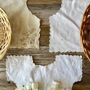 May include: An overhead shot features vintage embroidered linens with scalloped edges. Two pieces are cream-colored, one with "Hot Rolls" embroidered on it. Two white linens are also visible, one with a floral arrangement. Wicker baskets frame the scene.