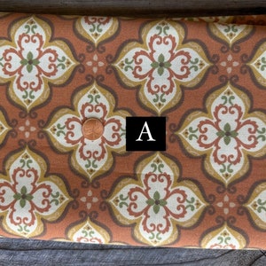 May include: Close-up of a fabric with a repeating floral pattern in shades of rust, cream, olive green, and gold. The design features stylized quatrefoils. A penny is placed on the fabric. The letter "A" is in a black square.