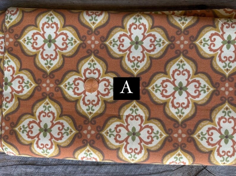 May include: Close-up of a fabric with a repeating floral pattern in shades of rust, cream, olive green, and gold. The design features stylized quatrefoils. A penny is placed on the fabric. The letter "A" is in a black square.