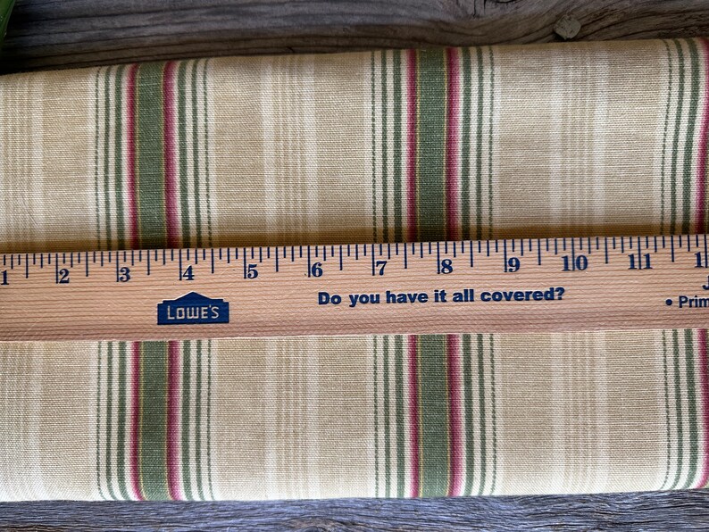 May include: Beige fabric with green and burgundy vertical stripes. A wooden ruler with the text "Do you have it all covered?" and the Lowe's logo is placed across the fabric. The ruler measures up to 28 cm.