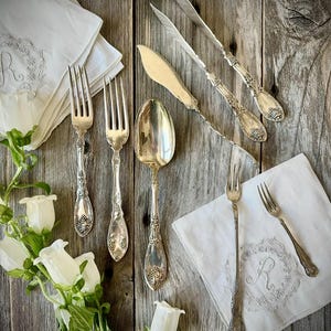 May include: A collection of silver-toned flatware including two dinner forks, one large spoon, two knives, and two smaller forks, arranged alongside two white embroidered napkins featuring the letter R and several white bell-shaped flowers.