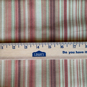 May include: Fabric with vertical stripes in beige, pink, and brown. A wooden ruler, marked in inches, lies across the fabric. The ruler has the Lowe's logo and the text "Do you have it all covered?" The ruler is approximately 23 cm long.
