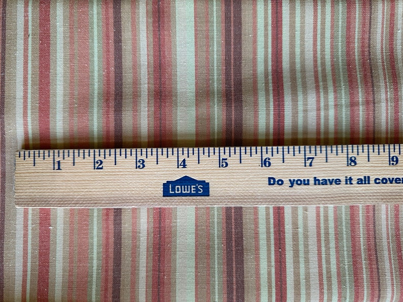 May include: Fabric with vertical stripes in beige, pink, and brown. A wooden ruler, marked in inches, lies across the fabric. The ruler has the Lowe's logo and the text "Do you have it all covered?" The ruler is approximately 23 cm long.